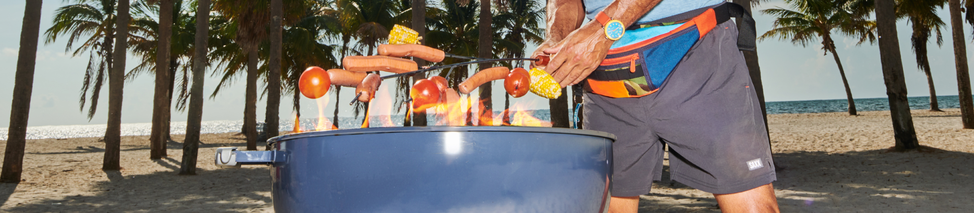 Man outside on beach with palm trees roasting hot dogs and corn over barbecue