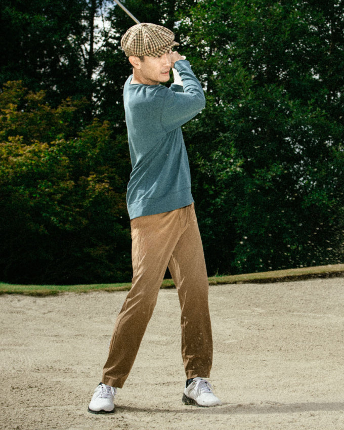 A man playing golf. He is mid swing and wearing khaki pants and a baker boy cap.