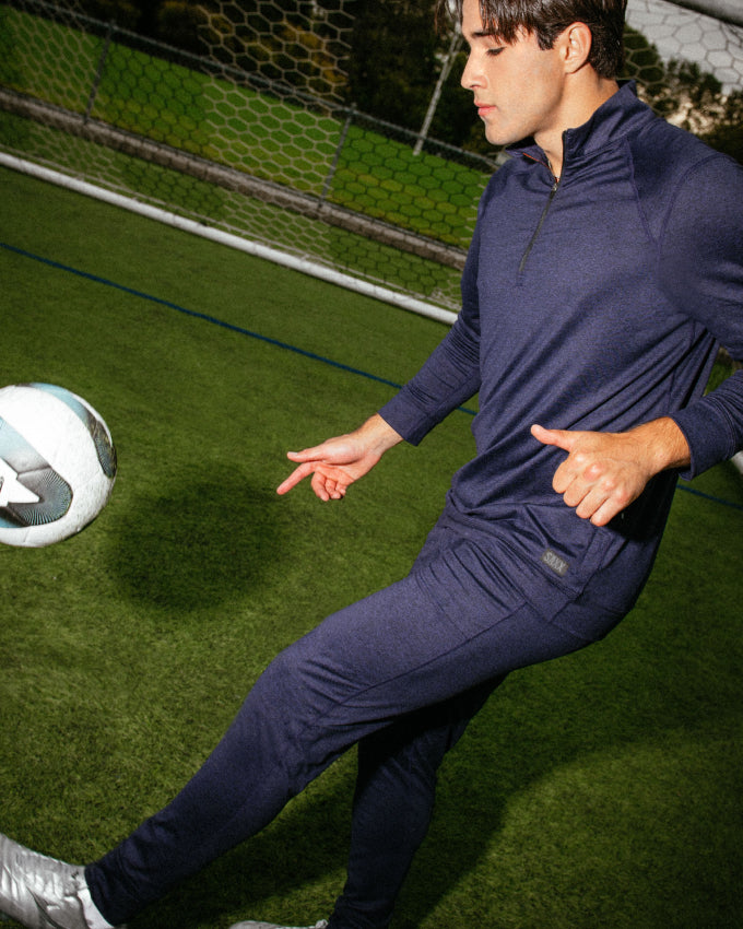 A man wearing a navy tracksuit doing kick ups on a soccer pitch.