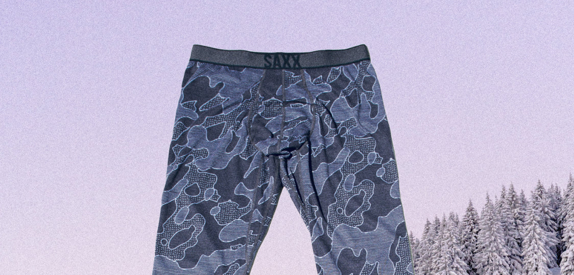 An image of a pair of long boxer briefs laid flat against a background that has snow covered pine trees and a vast sky. 