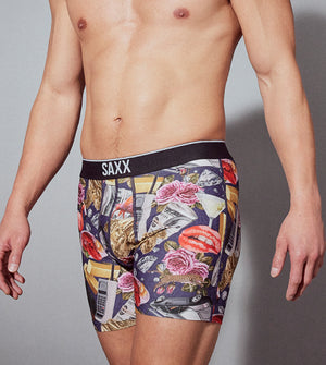 A man wearing SAXX boxer briefs with a multi-colored print including pink flowers, red lips, a martini class, and an old cell phone, posing on a neutral backdrop.