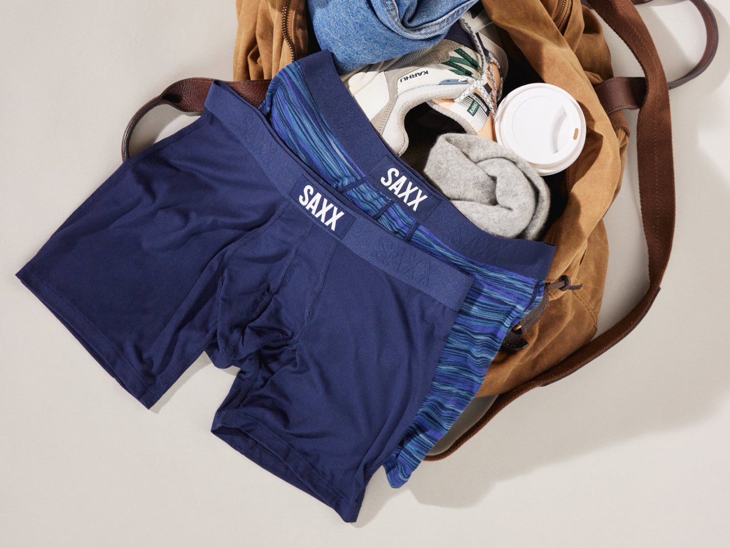 Two pairs of blue boxer briefs on top of a beige gym back which is open, revealing a sneaker and coffee cup.