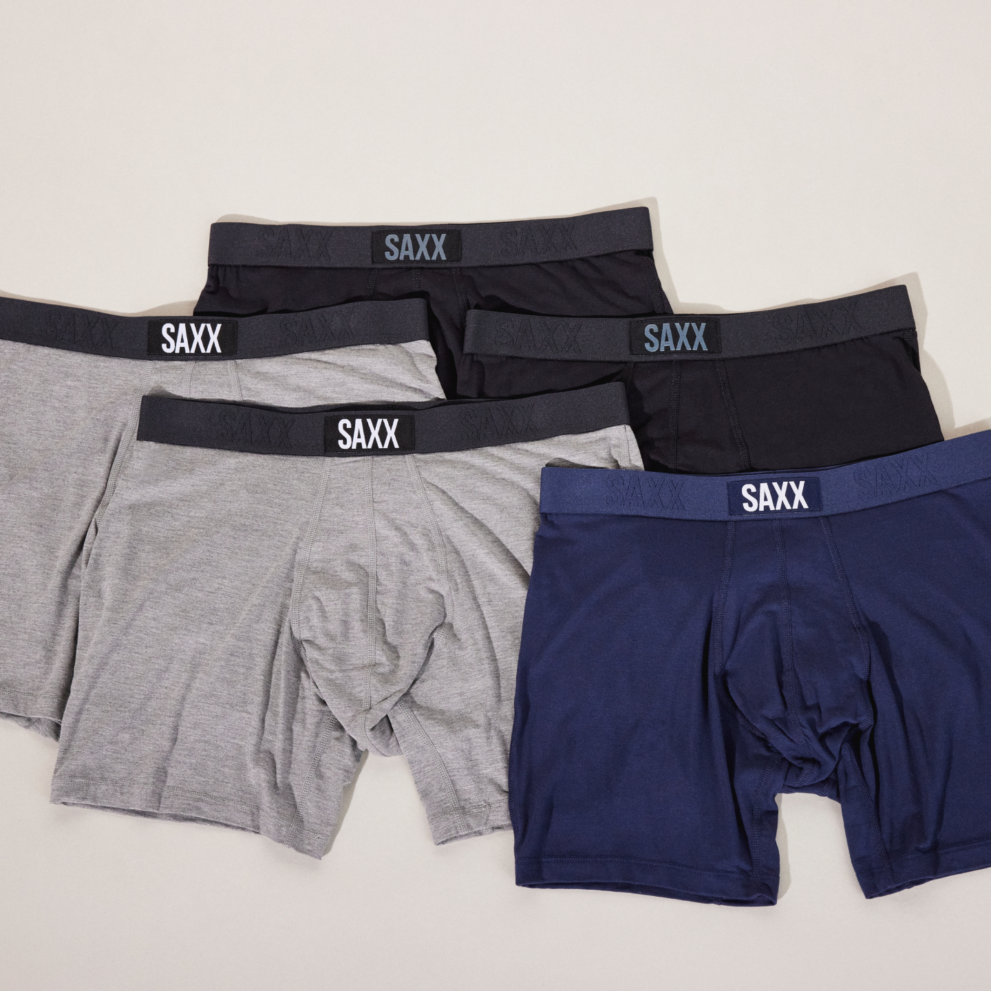 Five pairs of SAXX boxer briefs, two grey, two black, and one blue, laid out neatly against a neutral background. 