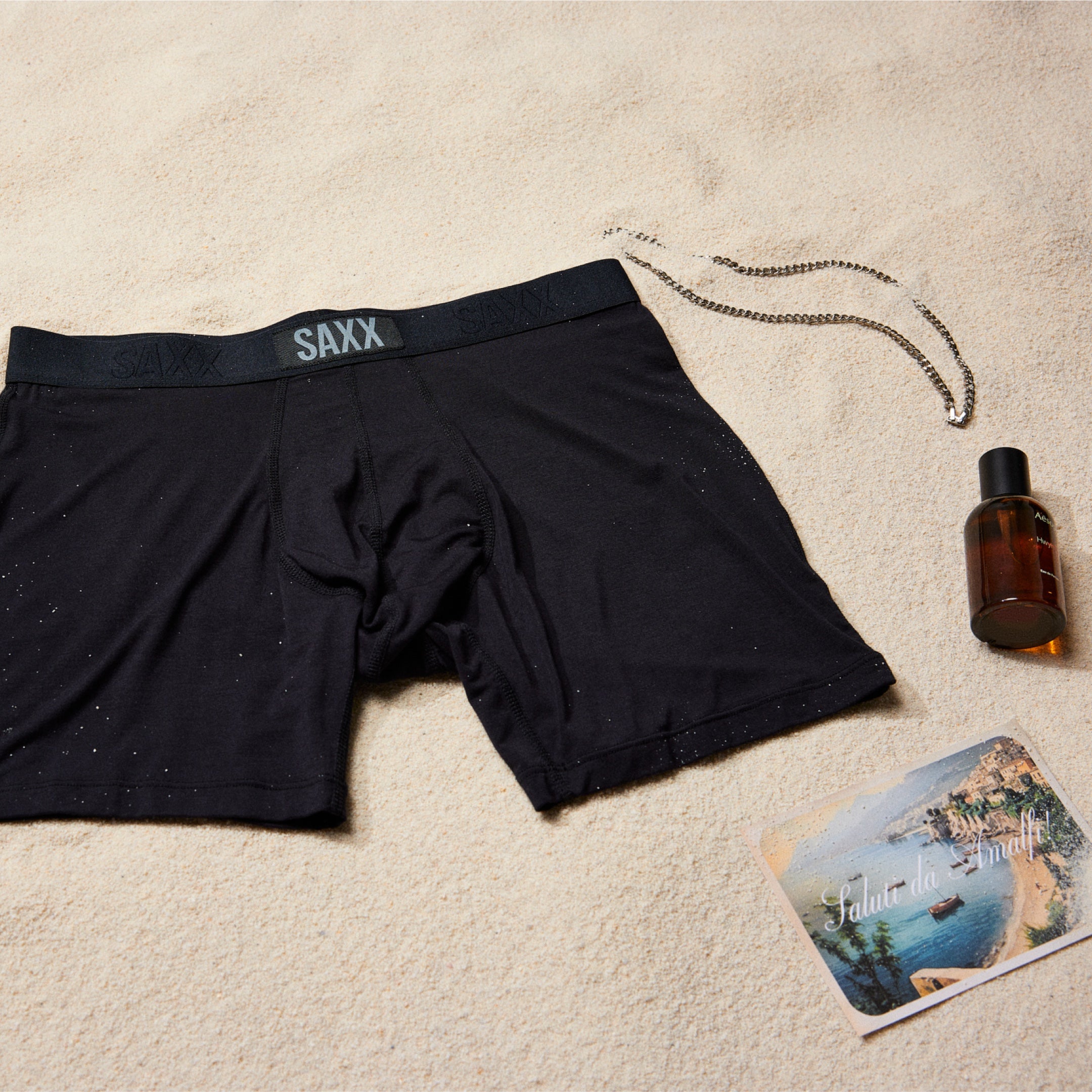 Black SAXX Boxer Briefs laid flat on the sand surrounded by a silver necklace, a sunscreen bottle, and a postcard.