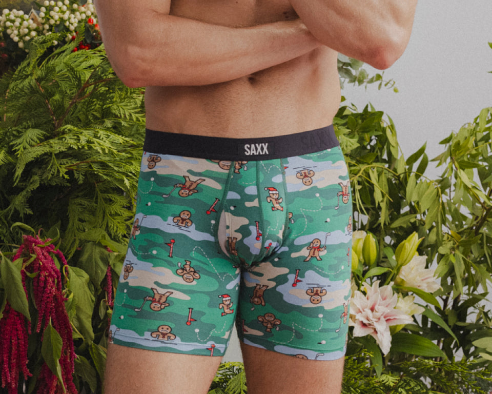 A close up of a man's underwear with a gingerbread man playing golf printed on them. He stands in front of foliage.