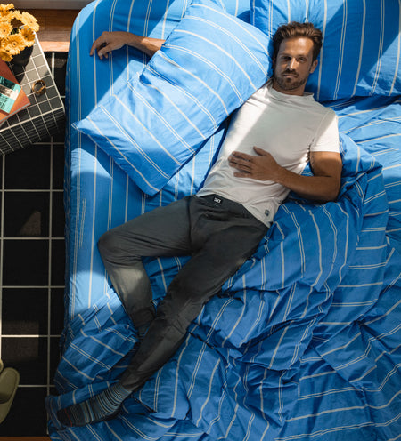 A man laying on a bed wearing a white t-shirt and grey pants seen from an aerial point of view directly overhead. 