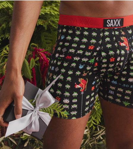 A man wearing black holiday-printed SAXX boxer briefs with a red band, appearing waist down, holding a grey gift box.