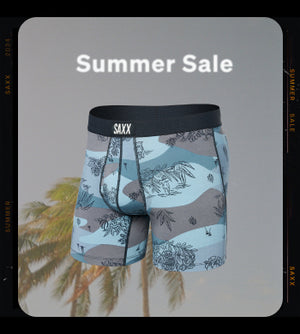 A snapshot of a swaying palm tree over blue sky with a pair of blue and grey SAXX boxer shorts with a tropical plant pattern overlaying the image along with the text "Summer Sale".  