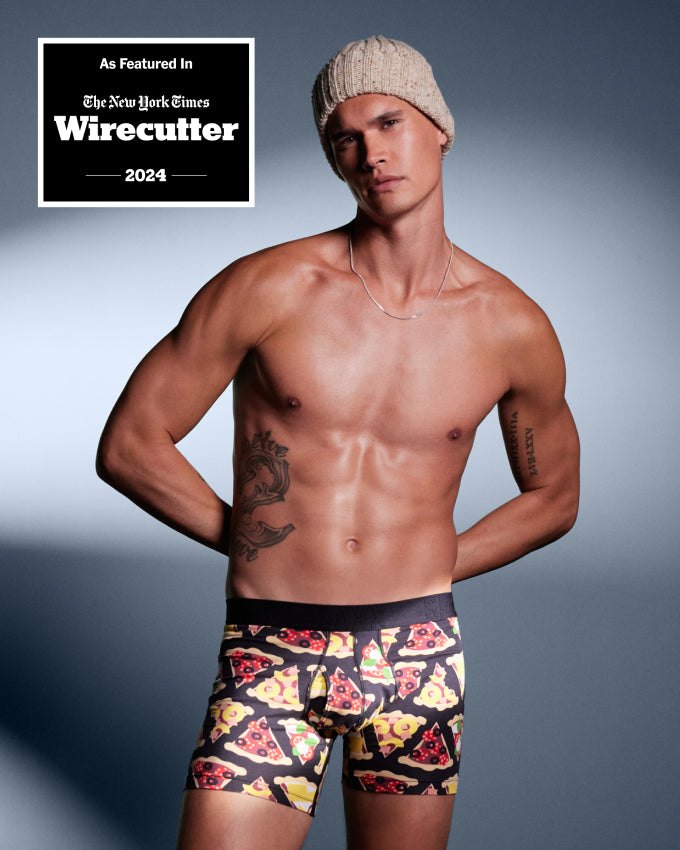 A man wearing black SAXX DropTemp® Cooling Cotton Boxer Briefs with a pizza print, and a cream-colored beanie, standing casually against a neutral backdrop with hands behind back. 