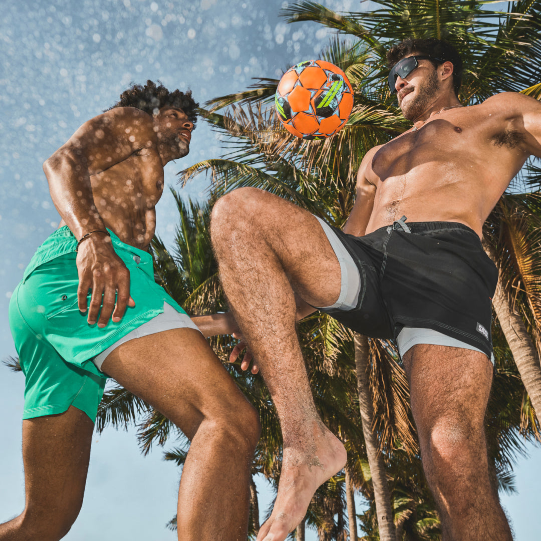 Man in green SAXX Swim shorts and man in black SAXX Swim shorts playing soccer at the beach, palm trees behind them.