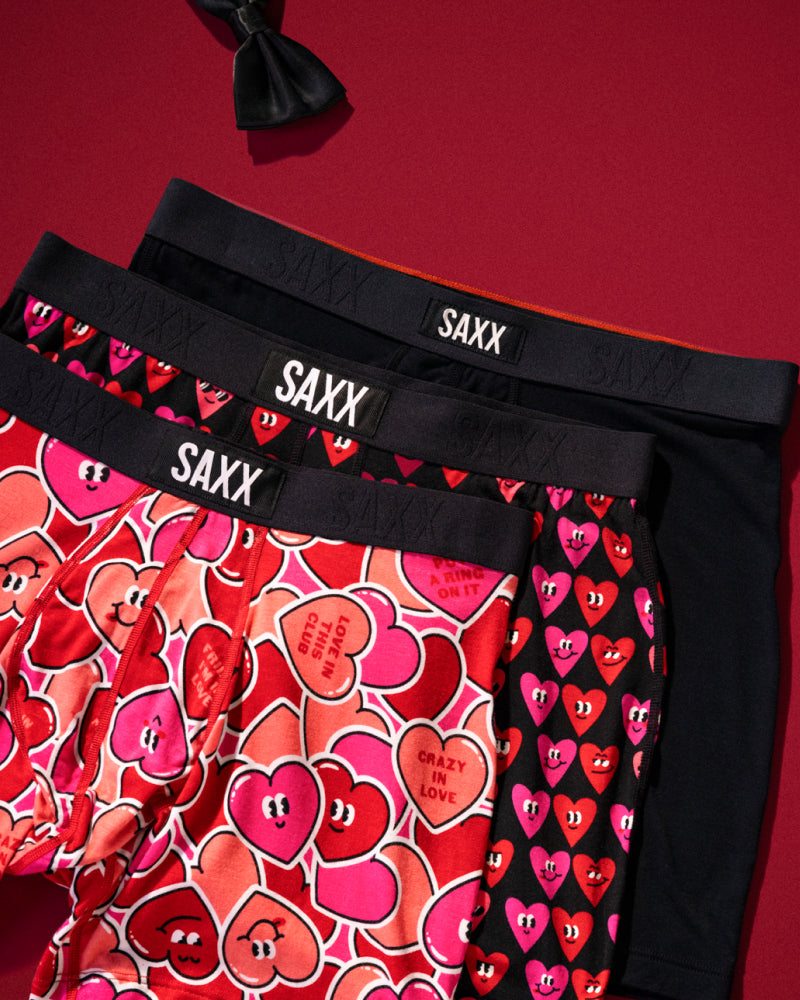 Three pairs of Valentine’s Day themed SAXX Boxer Briefs laid out flat over red background, black bowtie visible top left.