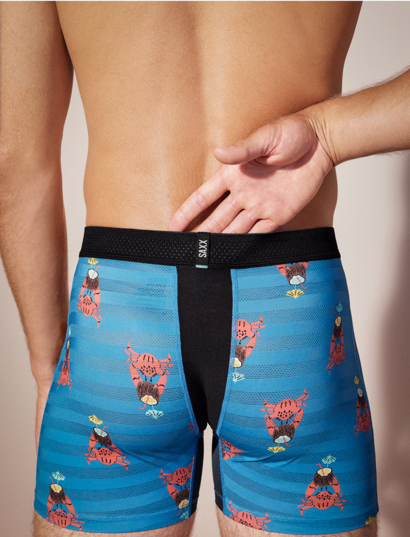 Closeup on backside of man wearing blue striped SAXX Mesh Boxer Briefs with tropical print, one hand in waistband.