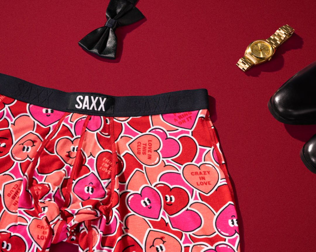 Valentine-themed SAXX Boxer Briefs with smiling candy hearts laid flat near bowtie, watch, and dress shoes, over red backdrop.