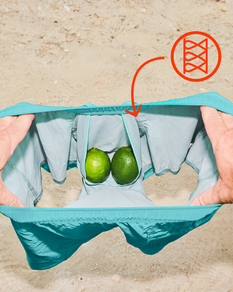 Hands holding open green SAXX Swim shorts, two limes in the BallPark Pouch® liner, icon indicating chafe-free Flat Out Seams®.