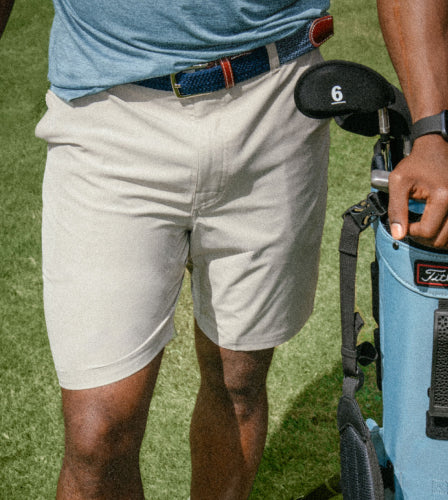 A man on a golf green with a golf bag and clubs wearing khaki SAXX 2N1 Shorts and a light blue SAXX top. 