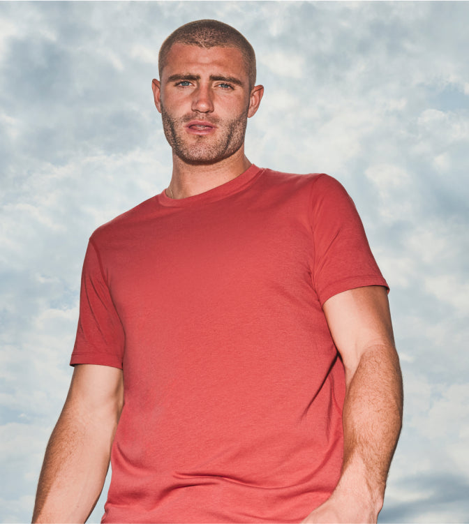 Man looking directly at camera wearing red SAXX tee in front of cloudy skies.