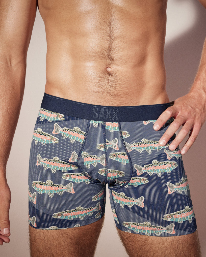 Closeup on male model wearing grey SAXX Boxer Briefs with fish pattern, one arm on hip, in front of neutral backdrop.