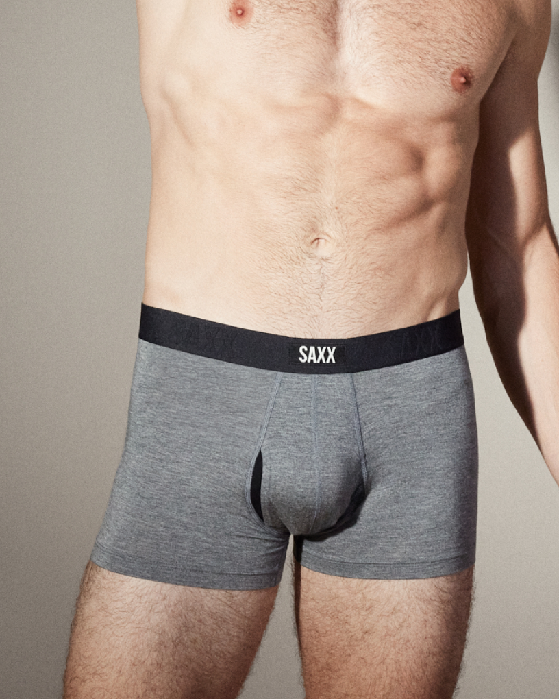 A fit man posing with one arm behind his head against a neutral backdrop wearing grey SAXX Trunks.