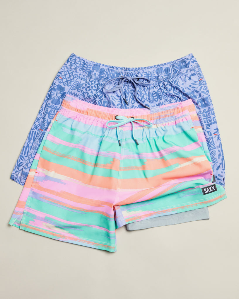 Two pairs of SAXX 2N1 Swim shorts, one patterned blue and one green with pink, laid out flat over a neutral backdrop.