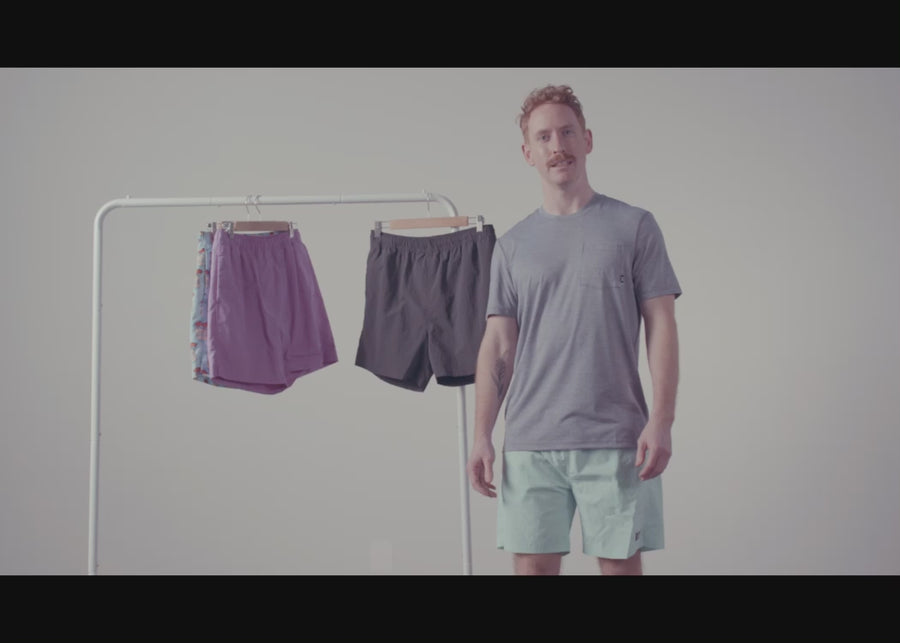 A man's hands showing the liner built into a pair of grey SAXX swim shorts hanging on a rack next to two other pairs.