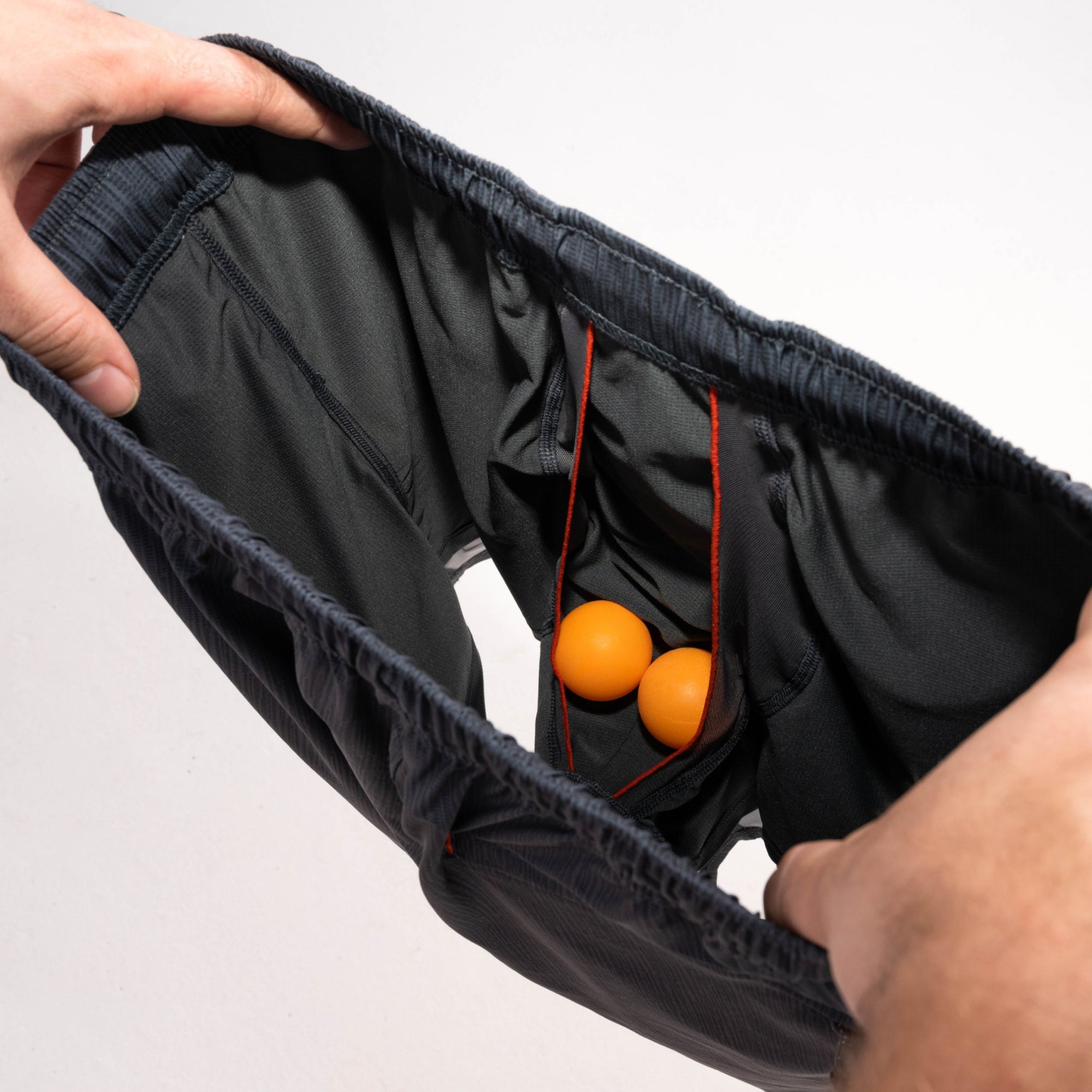 Man's hands revealing inside of 2N1 shorts showing liner with pouch holding two ping pong balls