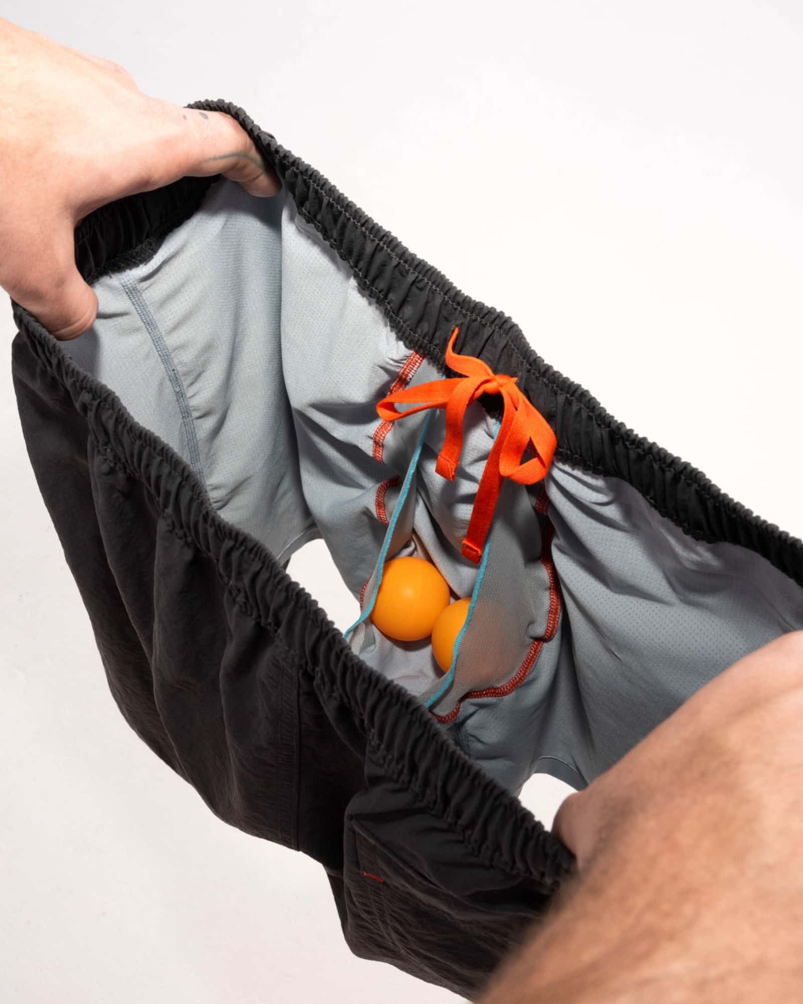 Man's hands revealing inside of 2N1 shorts showing liner with pouch holding two ping pong balls